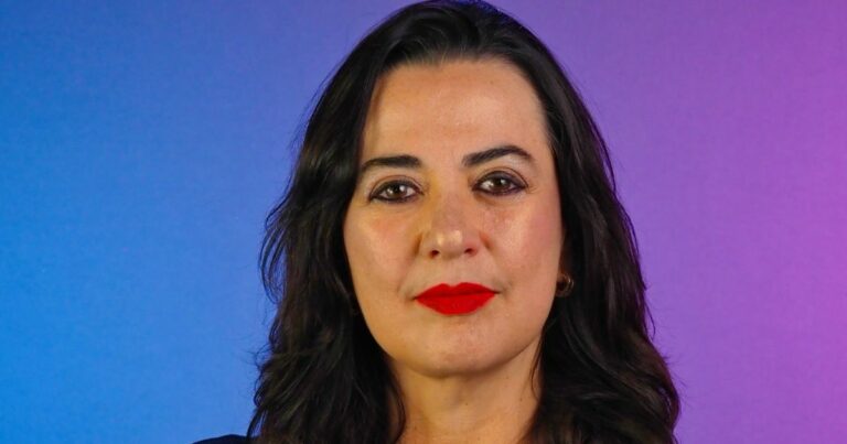 Retrato em close de uma pessoa posicionada de frente para a câmera. A pessoa tem cabelo escuro, liso e solto até abaixo dos ombros, e usa batom em tom vermelho intenso. O enquadramento mostra o rosto e parte superior do tronco. O fundo é liso com um degradê de cores, variando do azul à esquerda ao roxo à direita. A iluminação é uniforme e frontal, destacando os traços do rosto de maneira equilibrada, com aparência profissional e estúdio controlado. (Quatrix)