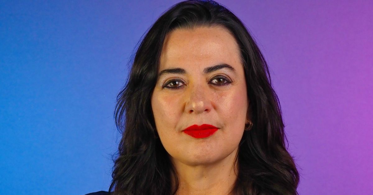 Retrato em close de uma pessoa posicionada de frente para a câmera. A pessoa tem cabelo escuro, liso e solto até abaixo dos ombros, e usa batom em tom vermelho intenso. O enquadramento mostra o rosto e parte superior do tronco. O fundo é liso com um degradê de cores, variando do azul à esquerda ao roxo à direita. A iluminação é uniforme e frontal, destacando os traços do rosto de maneira equilibrada, com aparência profissional e estúdio controlado. (Quatrix)