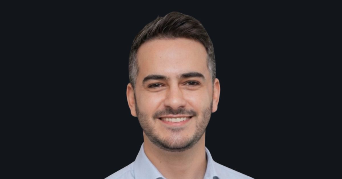 Retrato em close de uma pessoa posicionada de frente para a câmera. A pessoa tem cabelo curto escuro com laterais mais curtas e barba curta bem aparada, vestindo camisa clara com colarinho. O enquadramento mostra o rosto e parte superior dos ombros. O fundo é liso e escuro, sem elementos visuais adicionais, criando alto contraste com o rosto. A iluminação é uniforme e frontal, resultando em um retrato com aparência profissional e neutra. (Inter)
