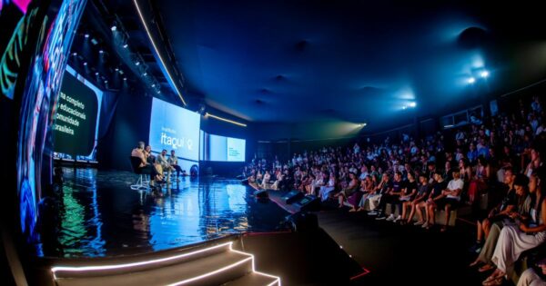 Imagem ampla de um palco em um evento corporativo ou tecnológico, visto a partir da lateral da plateia. No palco, várias pessoas estão sentadas em cadeiras individuais, participando de um painel de discussão. O piso do palco é escuro e altamente reflexivo, com degraus iluminados por faixas de luz branca na lateral frontal. Ao fundo, grandes telas digitais exibem conteúdos visuais e textos; em uma delas aparece o logotipo “itaqui Q” em letras claras sobre fundo escuro. À direita da imagem, uma plateia grande está sentada em arquibancadas, assistindo ao painel. O ambiente é fechado, com teto alto, iluminação azulada e estrutura técnica visível, reforçando o contexto de conferência formal voltada à tecnologia e inovação.