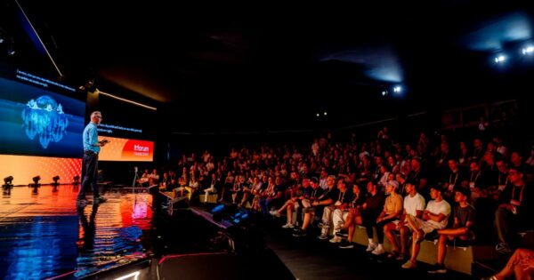 Imagem panorâmica de um auditório durante um evento corporativo ou tecnológico. À esquerda, um palco iluminado com piso escuro e reflexivo ocupa parte da cena. Sobre o palco, uma pessoa está em pé apresentando, voltada para uma plateia numerosa sentada em arquibancadas à direita da imagem. Ao fundo do palco, uma grande tela digital exibe gráficos visuais em tons de azul, semelhantes a uma representação de dados ou um globo estilizado. Na lateral da tela, há um painel laranja com o texto “itforum TRANCOSO”. A iluminação é cênica, com foco no palco, enquanto a plateia permanece em luz mais baixa. O ambiente é fechado, com teto escuro e estrutura de iluminação profissional.