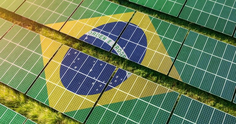 Imagem aérea de um grande conjunto de painéis solares dispostos em fileiras paralelas sobre o solo. Os painéis formam a imagem da bandeira do Brasil, com um losango amarelo, um círculo azul central e detalhes brancos, integrados ao padrão dos módulos fotovoltaicos. Entre as fileiras de painéis, é possível ver faixas de vegetação verde. As cores predominantes são verde, amarelo e azul, criando um contraste visual marcante. A composição associa energia solar e tecnologia à identidade visual brasileira, em um cenário externo bem iluminado por luz natural. (brasileiro)
