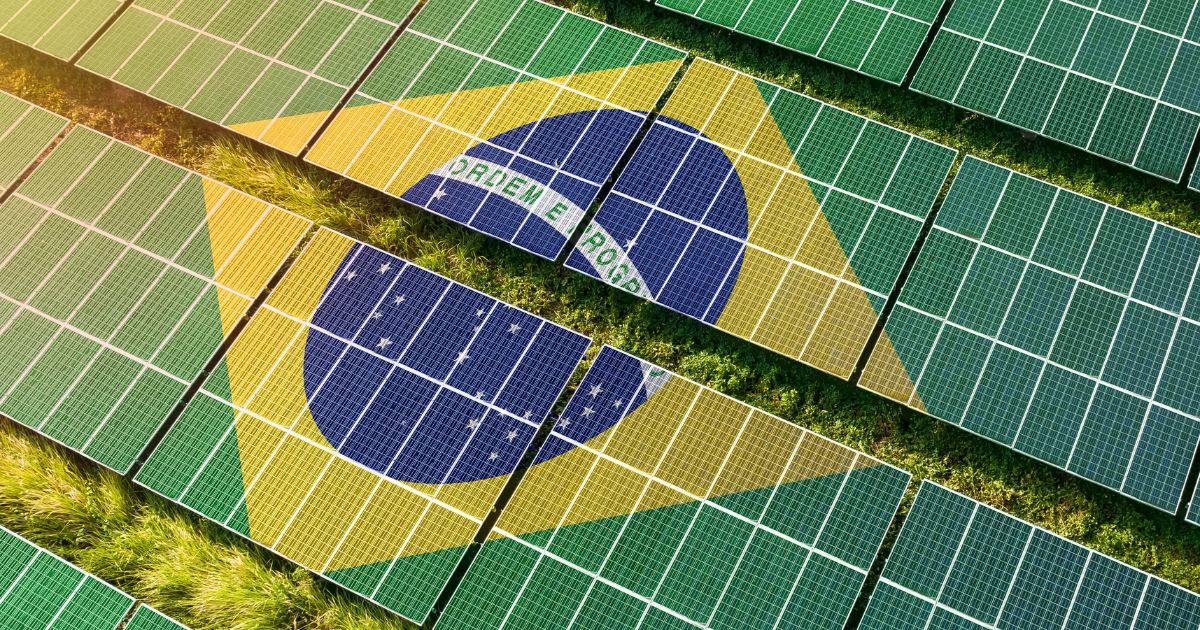 Imagem aérea de um grande conjunto de painéis solares dispostos em fileiras paralelas sobre o solo. Os painéis formam a imagem da bandeira do Brasil, com um losango amarelo, um círculo azul central e detalhes brancos, integrados ao padrão dos módulos fotovoltaicos. Entre as fileiras de painéis, é possível ver faixas de vegetação verde. As cores predominantes são verde, amarelo e azul, criando um contraste visual marcante. A composição associa energia solar e tecnologia à identidade visual brasileira, em um cenário externo bem iluminado por luz natural. (brasileiro)