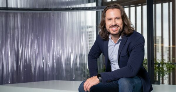 Pessoa sentada em ambiente interno moderno, inclinada levemente para frente e sorrindo para a câmera. Veste blazer azul escuro sobre camisa clara e jeans. Tem cabelo médio, liso e solto até os ombros, além de barba curta. O enquadramento mostra o corpo da cintura para cima, com um braço apoiado sobre a perna. Ao fundo, há uma divisória translúcida e grandes janelas com vista parcial de prédios e plantas, criando um ambiente corporativo contemporâneo bem iluminado por luz natural. (Daniel Campos, CHRO do Sem Parar)