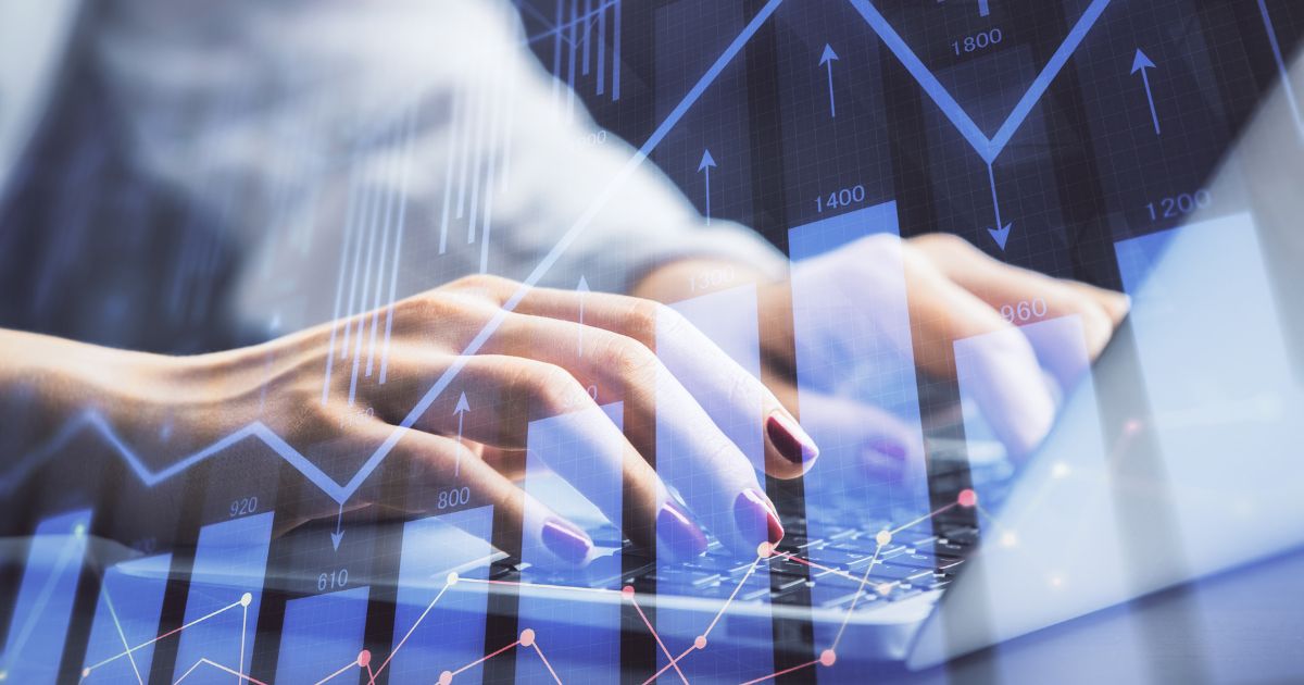 Imagem em close das mãos de uma pessoa digitando em um teclado de laptop. As unhas estão pintadas de vermelho escuro. Sobre a imagem, há uma sobreposição de gráficos financeiros digitais em tons de azul, com linhas, setas e barras apontando para cima, além de números e marcações, representando crescimento e análise de dados. A composição sugere atividades relacionadas a finanças, mercado de capitais, análise de desempenho ou ciência de dados em ambiente tecnológico. (IA agêntica, ciência)