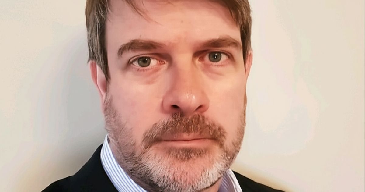 Retrato em close de uma pessoa posicionada de frente para a câmera, com expressão neutra e olhar direto. A pessoa tem cabelo curto, levemente despenteado, e barba curta com fios grisalhos. Veste blazer escuro sobre camisa clara com colarinho listrado. O enquadramento mostra o rosto e parte superior dos ombros. O fundo é liso e claro, sem elementos visíveis, e a iluminação é uniforme, destacando os traços faciais de forma equilibrada. (Citrix Latam)