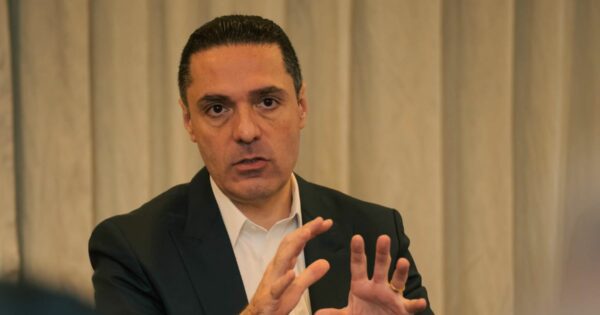 Pessoa em ambiente interno, posicionada de frente para a câmera, vestindo terno escuro sobre camisa branca. A pessoa tem cabelo curto e penteado para trás, sem barba, e apresenta expressão séria e concentrada. As mãos estão levantadas à frente do corpo, com os dedos levemente afastados, como se estivesse explicando ou enfatizando um ponto durante uma conversa ou apresentação. O fundo é composto por cortinas claras e homogêneas, desfocadas, criando um ambiente formal e profissional, com iluminação suave e uniforme. (Cisco)