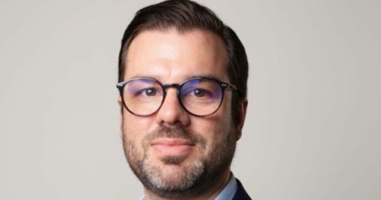 Retrato em close de uma pessoa posicionada de frente para a câmera, usando óculos de armação escura. A pessoa tem cabelo curto repartido para o lado e barba curta bem aparada. Veste blazer escuro sobre camisa clara. O enquadramento mostra o rosto e parte superior dos ombros. O fundo é liso e neutro em tom claro, sem elementos adicionais. A iluminação é uniforme, destacando os traços do rosto de forma equilibrada. (V8.Tech)