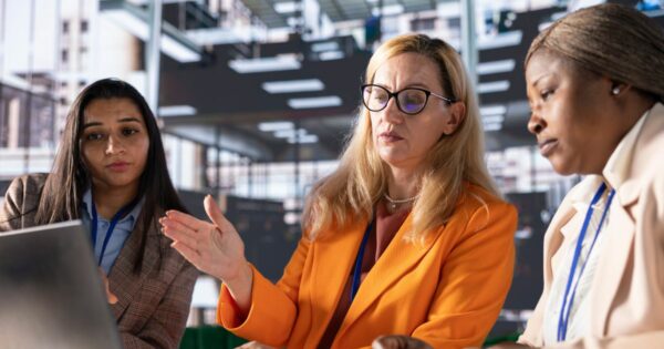 Imagem de três pessoas reunidas em um ambiente corporativo moderno, em frente a um laptop aberto. Ao centro, uma pessoa com cabelo claro e óculos, vestindo blazer laranja, gesticula com uma das mãos enquanto parece explicar algo. À esquerda, outra pessoa com cabelo escuro e longo observa atentamente a tela do laptop, vestindo blazer em tom neutro. À direita, uma pessoa com cabelo preso e vestindo blazer claro também observa a tela, com expressão concentrada. Todas usam crachás no pescoço. O fundo mostra um escritório amplo, com estrutura moderna e iluminação uniforme, sugerindo uma reunião ou colaboração profissional. (tecnologia)