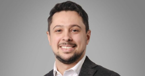 Retrato de uma pessoa posicionada de frente para a câmera, sorrindo. Tem cabelo curto escuro, barba curta e bem aparada e veste blazer escuro sobre camisa branca com colarinho. O enquadramento mostra o rosto e parte superior do tronco. O fundo é liso e cinza claro, sem elementos adicionais. A iluminação é uniforme e suave, destacando o rosto de forma equilibrada e criando um retrato com aparência profissional. (Genesys)