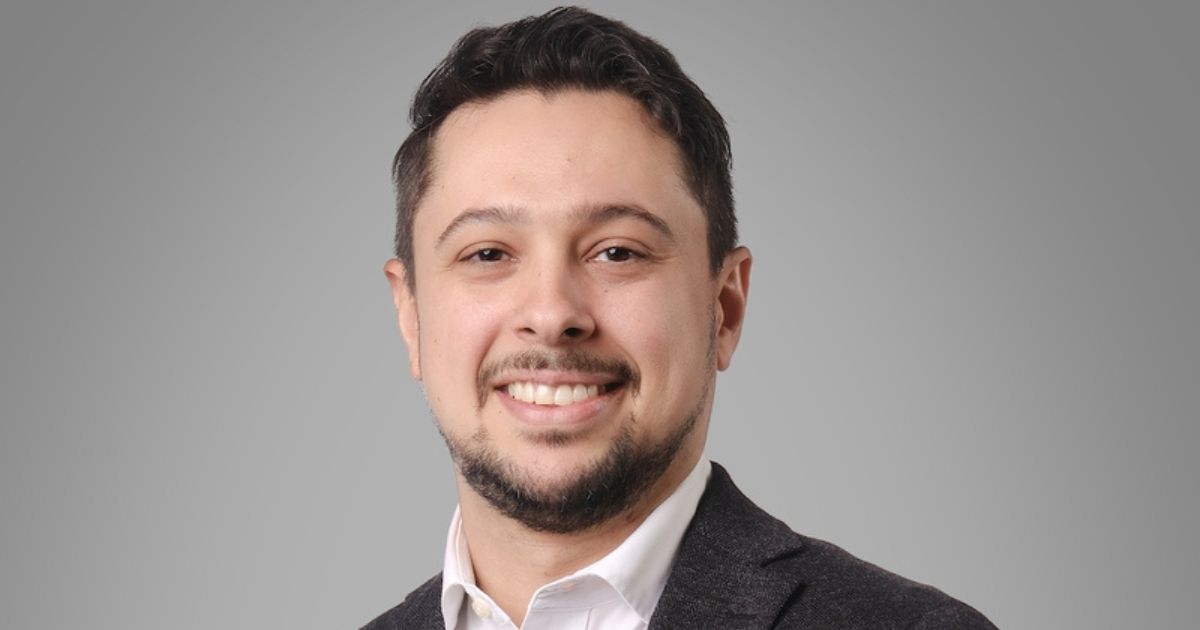 Retrato de uma pessoa posicionada de frente para a câmera, sorrindo. Tem cabelo curto escuro, barba curta e bem aparada e veste blazer escuro sobre camisa branca com colarinho. O enquadramento mostra o rosto e parte superior do tronco. O fundo é liso e cinza claro, sem elementos adicionais. A iluminação é uniforme e suave, destacando o rosto de forma equilibrada e criando um retrato com aparência profissional. (Genesys)