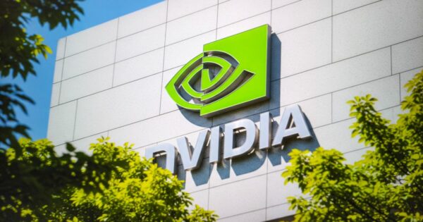 Imagem externa de um edifício corporativo moderno com fachada clara e revestimento em painéis retangulares. No centro da fachada, há o logotipo da NVIDIA: um símbolo verde em formato abstrato de olho, acompanhado do nome “NVIDIA” em letras metálicas tridimensionais. Árvores com folhas verdes aparecem em primeiro plano, parcialmente enquadrando o prédio, sugerindo um ambiente urbano arborizado. A iluminação natural indica um dia ensolarado, com céu azul ao fundo.