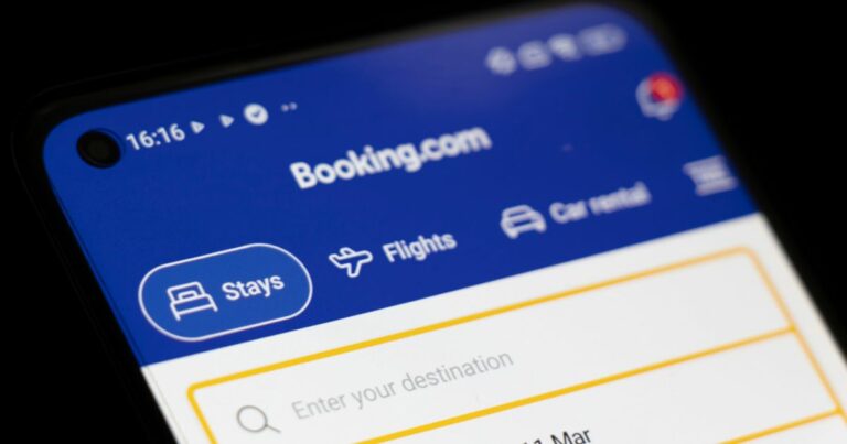 Imagem em close da tela de um smartphone exibindo o aplicativo Booking.com. A interface tem fundo azul na parte superior, com o logotipo “Booking.com” centralizado. Abaixo, aparecem opções de navegação com ícones e texto, como “Stays”, “Flights” e “Car rentals”. A opção “Stays” está destacada. Mais abaixo, há um campo de busca com o texto “Enter your destination”, delimitado por uma borda amarela. No canto superior esquerdo da tela, são visíveis ícones de status do dispositivo, incluindo horário, sinal e bateria. O fundo ao redor do celular é escuro, destacando a tela iluminada.