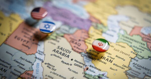 Mapa do Oriente Médio visto de perto, com foco na Arábia Saudita, Irã, Kuwait e Qatar. Sobre o mapa, há bottons redondos com bandeiras de diferentes países: um com a bandeira do Irã posicionado sobre o Irã, outro com a bandeira de Israel sobre a região próxima, e um com a bandeira dos Estados Unidos à esquerda. O fundo é o próprio mapa político, com nomes de países e cidades visíveis.