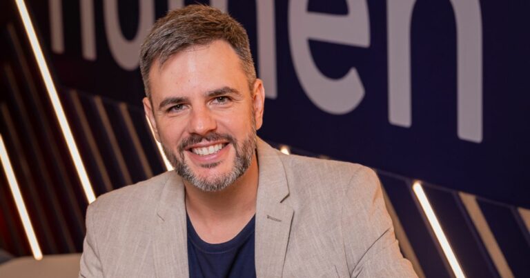 Retrato de uma pessoa adulta enquadrada do peito para cima, voltada para a câmera. A pessoa usa blazer claro sobre camiseta escura e tem cabelo curto com fios grisalhos e barba aparada. O fundo é escuro, com iluminação decorativa em linhas diagonais e letras grandes desfocadas, criando um ambiente moderno e corporativo. (Rafael Cabral)
