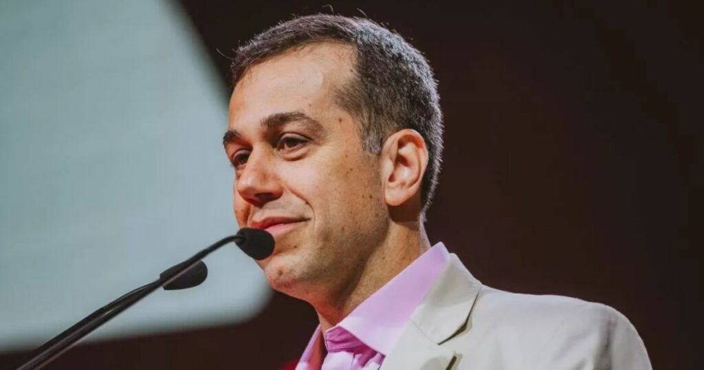 Retrato em close de uma pessoa posicionada atrás de um microfone em um púlpito, enquadrada do ombro para cima. A pessoa veste blazer claro sobre camisa em tom rosado e está inclinada levemente em direção ao microfone. Ao fundo, há um cenário com iluminação em tons quentes, predominantemente vermelho e laranja, criando contraste com o primeiro plano. A iluminação destaca o rosto e a parte superior do corpo, sugerindo um ambiente de palestra, evento ou apresentação. (TCS, Charles)