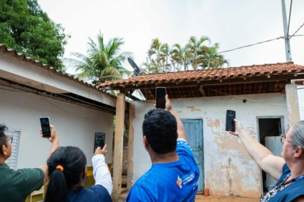 Grupo de pessoas em área residencial rural levantando celulares para captar sinal de internet em frente a casas simples, ilustrando desafios de conectividade e acesso digital em regiões afastadas do Brasil. (conectividade)