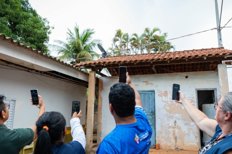 Grupo de pessoas em área residencial rural levantando celulares para captar sinal de internet em frente a casas simples, ilustrando desafios de conectividade e acesso digital em regiões afastadas do Brasil. (conectividade)