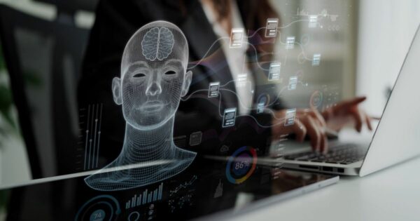 Pessoa utilizando um computador portátil em uma mesa, vista parcialmente ao fundo. Em primeiro plano, há uma sobreposição gráfica translúcida com a representação de uma cabeça humana em estilo wireframe, mostrando o cérebro em destaque. Ao redor da figura aparecem painéis digitais flutuantes com ícones, gráficos circulares, barras, porcentagens e blocos conectados por linhas, sugerindo processamento de dados e sistemas digitais. O ambiente tem iluminação suave, com fundo desfocado que mantém o foco nos elementos tecnológicos. (Brasil)