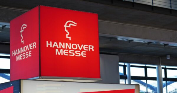 Estrutura cúbica suspensa em ambiente interno exibe a marca ‘HANNOVER MESSE’ em letras brancas sobre fundo vermelho intenso, acompanhada de um logotipo estilizado em formato de perfil. O cubo está posicionado acima de um estande, dentro de um pavilhão com teto alto, vigas metálicas e janelas amplas que deixam entrar luz natural. O ambiente sugere uma feira ou evento industrial e tecnológico.