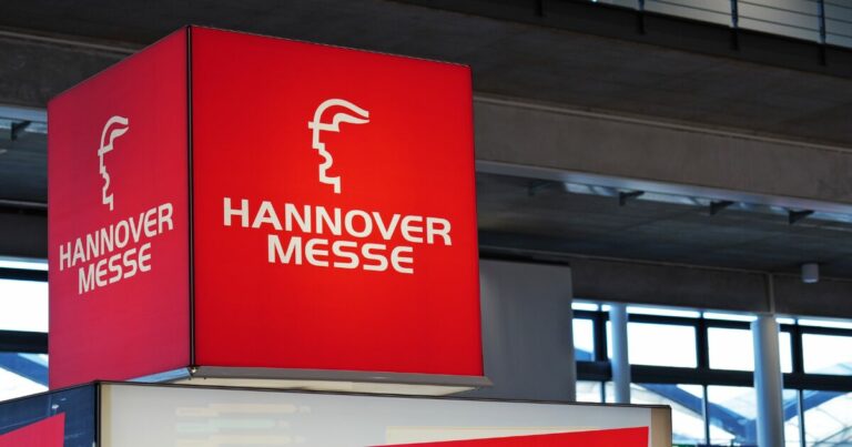 Estrutura cúbica suspensa em ambiente interno exibe a marca ‘HANNOVER MESSE’ em letras brancas sobre fundo vermelho intenso, acompanhada de um logotipo estilizado em formato de perfil. O cubo está posicionado acima de um estande, dentro de um pavilhão com teto alto, vigas metálicas e janelas amplas que deixam entrar luz natural. O ambiente sugere uma feira ou evento industrial e tecnológico.