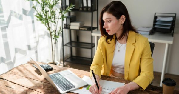 Pessoa adulta sentada à mesa de madeira em um ambiente interno bem iluminado, escrevendo em papéis com uma caneta enquanto utiliza um computador portátil aberto à frente. A pessoa veste blazer amarelo sobre uma blusa clara. Sobre a mesa há documentos e um copo descartável. Ao fundo, há uma estante com pastas e livros, uma planta em vaso próxima à janela com cortina clara e iluminação natural entrando pela lateral, criando um ambiente de trabalho organizado e moderno. (empreendedores)