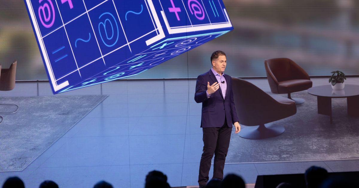 Pessoa em pé sobre um palco durante uma apresentação, enquadrada de corpo inteiro, usando blazer escuro sobre camisa clara e microfone de lapela. A pessoa gesticula com uma das mãos enquanto fala. Ao fundo, há uma grande tela exibindo um ambiente com poltronas e uma mesa redonda, além de um grande cubo gráfico suspenso com símbolos e números coloridos. Na parte inferior da imagem, aparecem silhuetas desfocadas da plateia. A iluminação é direcionada ao palco, com fundo levemente azulado, sugerindo um evento corporativo ou conferência. (Dell)