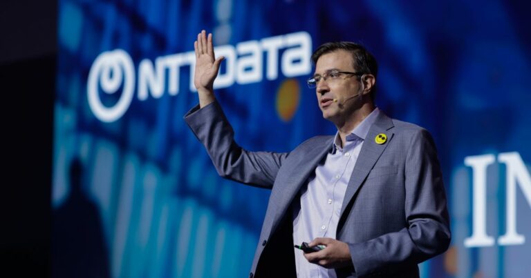 Pessoa em pé durante uma apresentação, enquadrada do tronco para cima, vestindo blazer cinza sobre camisa clara e usando óculos. A pessoa levanta uma das mãos enquanto segura um pequeno controle na outra. Ao fundo, há um grande painel iluminado em tons de azul com o logotipo ‘NTT DATA’ visível. A iluminação é direcionada para a pessoa, destacando-a em contraste com o fundo e sugerindo um evento corporativo ou tecnológico.