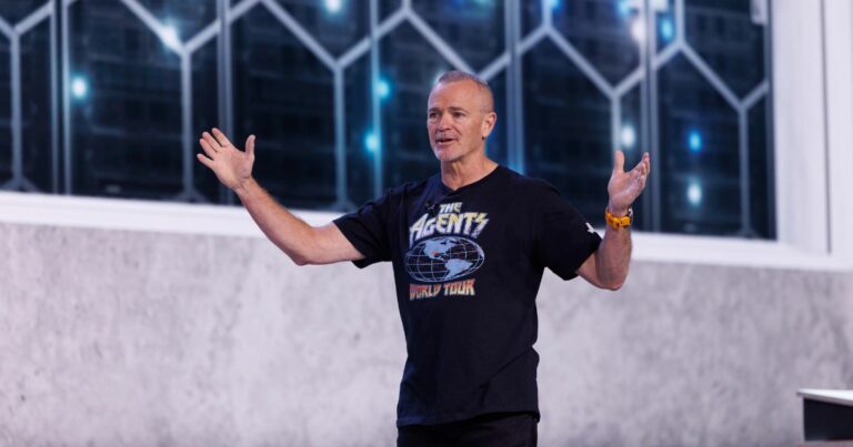Pessoa em pé em um palco durante uma apresentação, enquadrada do corpo até a cabeça. A pessoa veste camiseta escura com estampa visível contendo o texto ‘The Agents World Tour’. Os braços estão abertos para os lados, com as mãos levantadas, em posição de fala. Ao fundo, há uma parede clara na parte inferior e uma área superior com painéis de vidro em formato geométrico, com luzes azuis visíveis através deles. A iluminação é direcionada para a pessoa, destacando-a em relação ao fundo, sugerindo um evento ou conferência de tecnologia. (Dell)