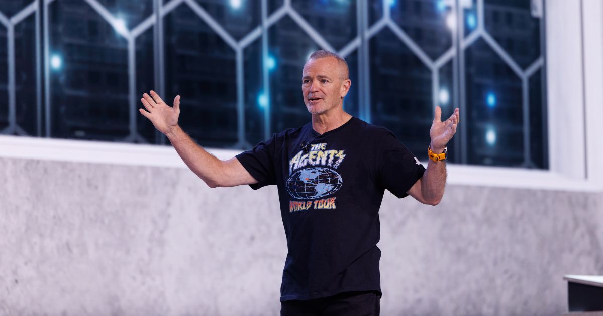 Pessoa em pé em um palco durante uma apresentação, enquadrada do corpo até a cabeça. A pessoa veste camiseta escura com estampa visível contendo o texto ‘The Agents World Tour’. Os braços estão abertos para os lados, com as mãos levantadas, em posição de fala. Ao fundo, há uma parede clara na parte inferior e uma área superior com painéis de vidro em formato geométrico, com luzes azuis visíveis através deles. A iluminação é direcionada para a pessoa, destacando-a em relação ao fundo, sugerindo um evento ou conferência de tecnologia. (Dell)