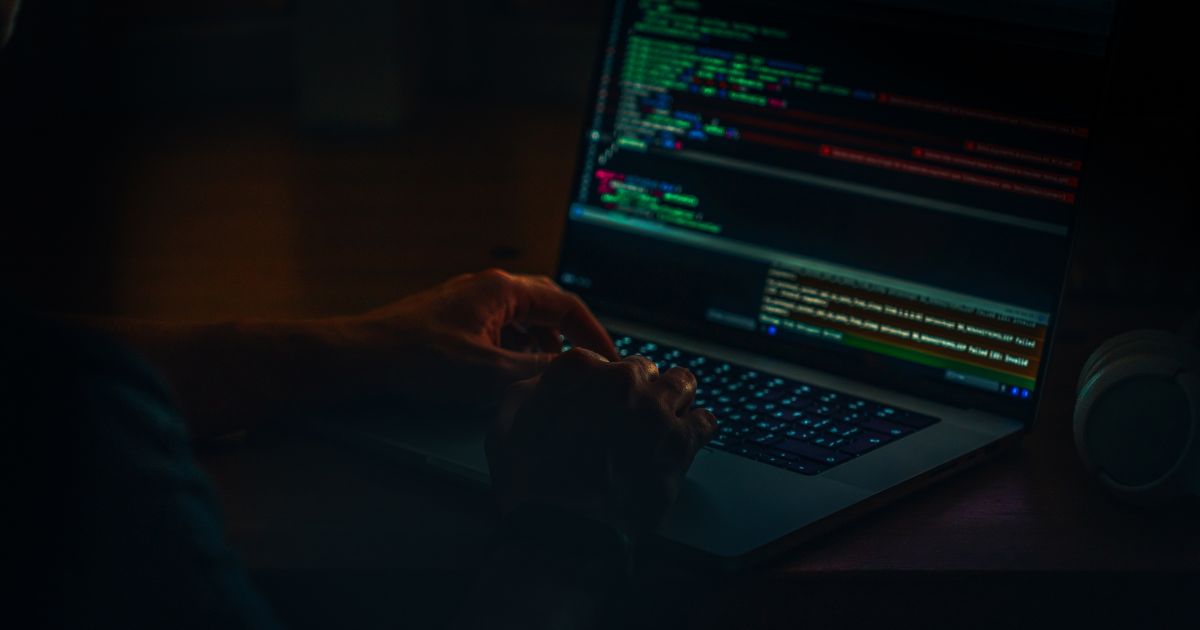 Ambiente escuro com uma pessoa utilizando um computador portátil sobre uma mesa. A tela do laptop exibe linhas de código coloridas em diferentes tons, como verde, vermelho e azul, organizadas em janelas de programação. As mãos da pessoa estão posicionadas sobre o teclado, em ação de digitação. À direita do computador, há um objeto cilíndrico claro, possivelmente um alto-falante ou recipiente. A iluminação é baixa, com o brilho da tela destacando as mãos e o teclado, criando um cenário associado a programação ou desenvolvimento de software.