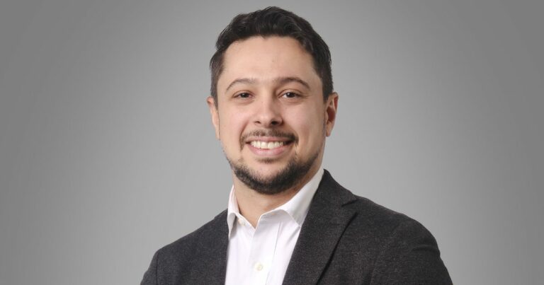 Retrato de uma pessoa adulta enquadrada do peito para cima, voltada para a câmera. A pessoa veste camisa branca com colarinho e blazer escuro. O cabelo é curto e o rosto apresenta barba curta. O fundo é liso em tom cinza claro, com iluminação uniforme que destaca o rosto e cria um visual neutro e profissional. (Genesys)