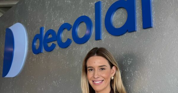 Pessoa adulta enquadrada do peito para cima, posicionada em frente a uma parede com o logotipo ‘decolar’ em letras azuis tridimensionais. A pessoa está parcialmente enquadrada à direita da imagem e olha em direção à câmera. O fundo é uma parede cinza texturizada, com boa iluminação, criando um cenário corporativo e profissional.