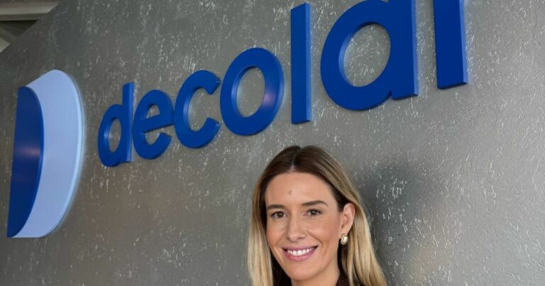 Pessoa adulta enquadrada do peito para cima, posicionada em frente a uma parede com o logotipo ‘decolar’ em letras azuis tridimensionais. A pessoa está parcialmente enquadrada à direita da imagem e olha em direção à câmera. O fundo é uma parede cinza texturizada, com boa iluminação, criando um cenário corporativo e profissional.