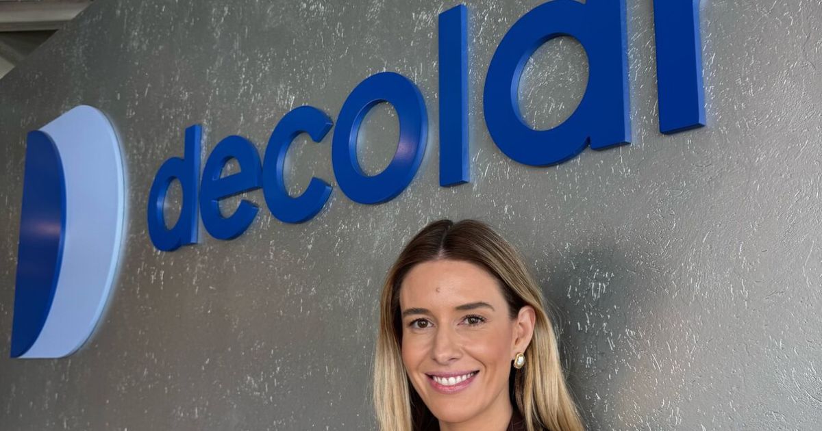 Pessoa adulta enquadrada do peito para cima, posicionada em frente a uma parede com o logotipo ‘decolar’ em letras azuis tridimensionais. A pessoa está parcialmente enquadrada à direita da imagem e olha em direção à câmera. O fundo é uma parede cinza texturizada, com boa iluminação, criando um cenário corporativo e profissional.