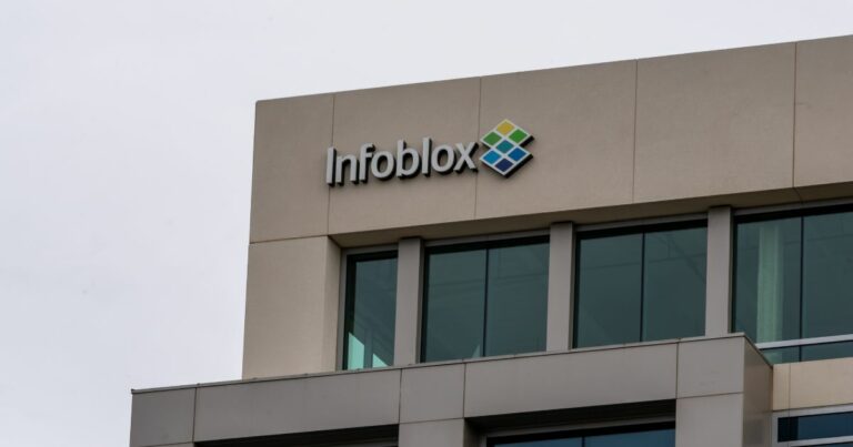 Parte superior da fachada de um edifício comercial visto de baixo, com janelas de vidro escuro e revestimento em tom bege. No topo da estrutura aparece o logotipo ‘Infoblox’ em letras pretas, acompanhado de um símbolo geométrico colorido à direita do nome. O céu ao fundo é claro e nublado, criando um contraste suave com o prédio.