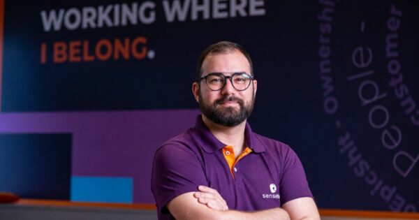 Pessoa adulta enquadrada do peito para cima, em posição frontal, com braços cruzados. A pessoa usa camisa polo em tom roxo com detalhes em laranja e óculos de armação escura. O fundo é um ambiente interno com paredes escuras e elementos gráficos desfocados. Ao fundo, é possível ler o texto ‘WORKING WHERE I BELONG.’ e outras palavras verticais parcialmente visíveis, criando um cenário corporativo e moderno. (Sensedia)