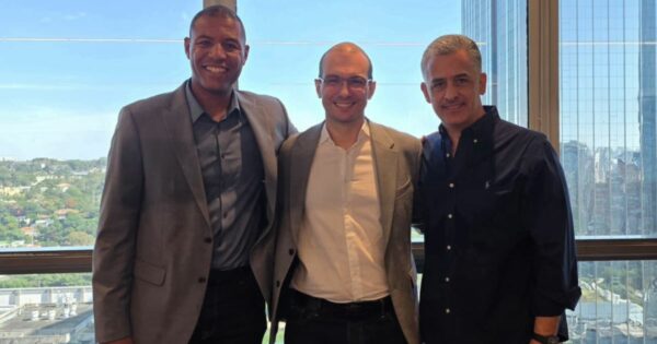 Três pessoas adultas em pé, lado a lado, olhando para a câmera, enquadradas do tronco para cima. As três vestem roupas de estilo corporativo, como blazers, camisas sociais e camisa de manga longa. O ambiente é interno, com uma grande janela ao fundo que mostra uma paisagem urbana com prédios e áreas verdes durante o dia. A iluminação é natural, vinda da janela, criando um cenário profissional e informal ao mesmo tempo. (Forlex)