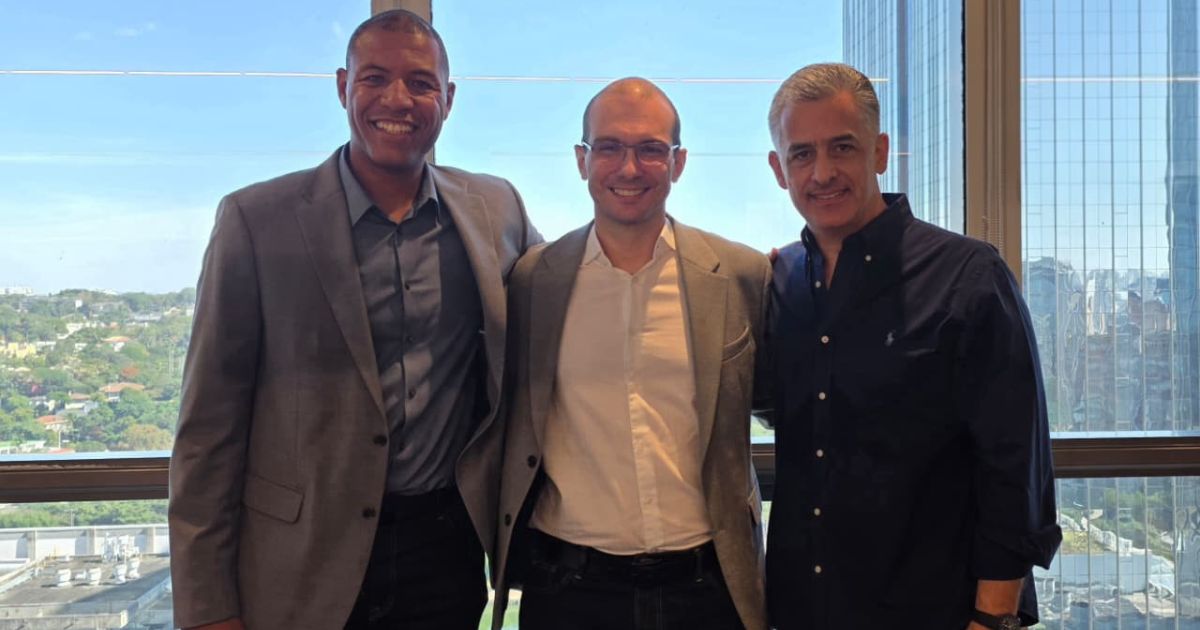 Três pessoas adultas em pé, lado a lado, olhando para a câmera, enquadradas do tronco para cima. As três vestem roupas de estilo corporativo, como blazers, camisas sociais e camisa de manga longa. O ambiente é interno, com uma grande janela ao fundo que mostra uma paisagem urbana com prédios e áreas verdes durante o dia. A iluminação é natural, vinda da janela, criando um cenário profissional e informal ao mesmo tempo. (Forlex)