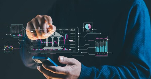 Mãos de uma pessoa segurando um smartphone enquanto a outra mão toca uma interface digital projetada à frente do dispositivo. No centro, há um painel com gráfico financeiro em formato de candlestick e a sigla ‘IT’. Ao redor do painel principal, aparecem outros gráficos e indicadores analíticos conectados por linhas, incluindo gráficos de barras, pizza e linhas em tons de azul, rosa e verde. A pessoa veste um casaco escuro com capuz. O fundo é escuro, com iluminação direcionada para as mãos e os elementos gráficos, sugerindo uso de tecnologia para análise de dados ou mercado financeiro. (transformação digital)
