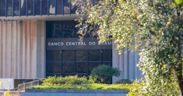 Fachada de um edifício institucional com colunas verticais claras e grandes janelas de vidro. No centro da imagem, acima da entrada, aparece o texto ‘BANCO CENTRAL DO BRASIL’. Em primeiro plano há um jardim com plantas e flores, e à direita parte da cena é enquadrada por uma árvore com folhas iluminadas pela luz natural. O ambiente é externo e bem iluminado.
