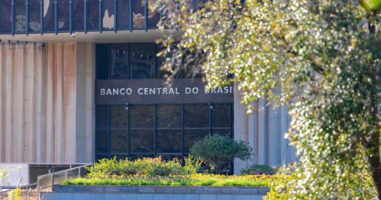 Fachada de um edifício institucional com colunas verticais claras e grandes janelas de vidro. No centro da imagem, acima da entrada, aparece o texto ‘BANCO CENTRAL DO BRASIL’. Em primeiro plano há um jardim com plantas e flores, e à direita parte da cena é enquadrada por uma árvore com folhas iluminadas pela luz natural. O ambiente é externo e bem iluminado.