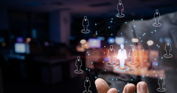 Mão de uma pessoa estendida em primeiro plano com um elemento gráfico digital sobreposto, formando uma rede de conexões. No centro da rede, há um ícone maior representando uma figura humana destacada, cercada por outros ícones menores distribuídos ao redor e conectados por linhas e pontos. O fundo é desfocado, com luzes e elementos de um ambiente interno, sugerindo um contexto tecnológico relacionado a redes profissionais, conexões ou gestão de pessoas. (qualidade)