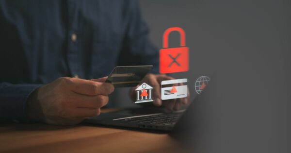 Mãos de uma pessoa segurando um cartão bancário enquanto utiliza um computador portátil apoiado sobre uma mesa. Sobre a imagem aparecem ícones digitais translúcidos, incluindo um cadeado vermelho com um símbolo de bloqueio, um ícone de alerta, um cartão com aviso e um símbolo institucional, sugerindo risco, bloqueio ou segurança em transações financeiras. O fundo é escuro e desfocado, reforçando um contexto de tecnologia e segurança digital. (ciber)
