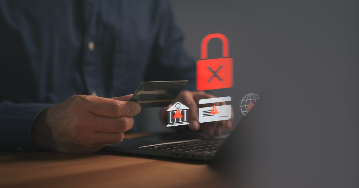 Mãos de uma pessoa segurando um cartão bancário enquanto utiliza um computador portátil apoiado sobre uma mesa. Sobre a imagem aparecem ícones digitais translúcidos, incluindo um cadeado vermelho com um símbolo de bloqueio, um ícone de alerta, um cartão com aviso e um símbolo institucional, sugerindo risco, bloqueio ou segurança em transações financeiras. O fundo é escuro e desfocado, reforçando um contexto de tecnologia e segurança digital. (ciber)