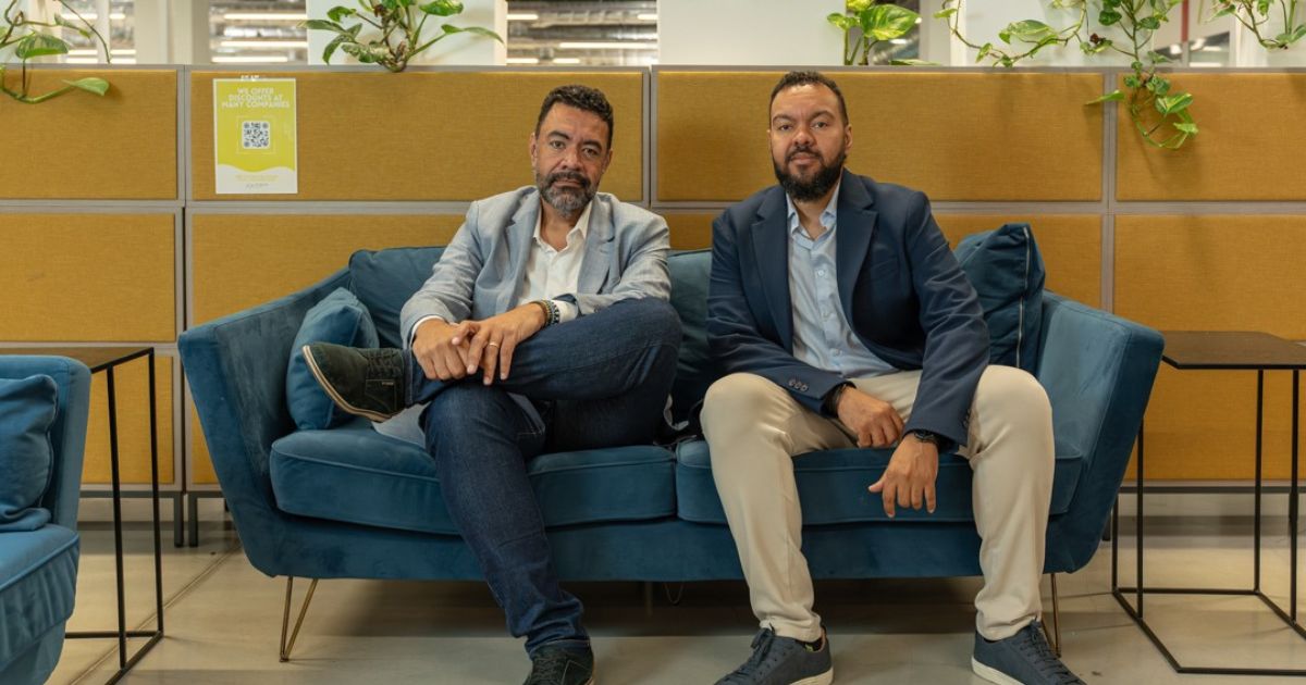 Imagem de um ambiente corporativo moderno com duas pessoas sentadas em um sofá azul em uma área de convivência. O espaço apresenta design contemporâneo, com painel amadeirado ao fundo, divisórias organizadas e plantas decorativas que trazem um toque natural ao escritório. A decoração inclui mesas laterais minimalistas e iluminação uniforme, criando um ambiente profissional, organizado e acolhedor, ideal para reuniões informais, networking ou momentos de pausa dentro do ambiente de trabalho. (AENVO)
