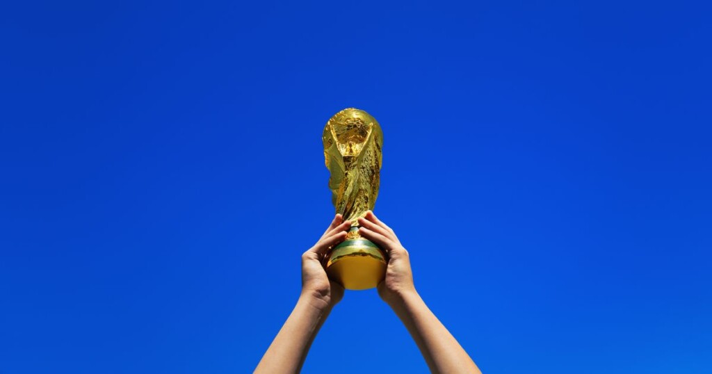 Fundo uniforme em azul intenso ocupando toda a cena. No centro inferior da imagem, duas mãos levantam um troféu dourado com formato de figura humana estilizada segurando um globo na parte superior. O objeto possui acabamento brilhante e refletivo. As mãos estão posicionadas por baixo da base do troféu, sustentando-o acima da linha do horizonte visual, criando destaque para o objeto contra o fundo liso e sem outros elementos. (Copa)