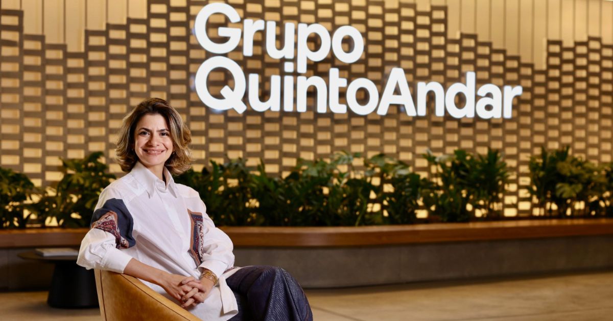 Pessoa sentada em poltrona em ambiente corporativo moderno, com painel decorativo ao fundo, plantas e logotipo “Grupo QuintoAndar” em destaque na parede.