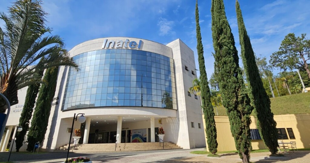 Imagem externa de um edifício institucional moderno, com fachada principal curva revestida por grandes painéis de vidro refletivo. No topo da estrutura, está visível o nome “Inatel” em letras grandes. A entrada principal fica no centro, acessada por uma escadaria e apoiada por colunas claras. Na área ao redor do prédio, há paisagismo com árvores altas e estreitas, incluindo ciprestes, além de uma palmeira no lado esquerdo da imagem. O céu está claro, com tons de azul e poucas nuvens, indicando iluminação natural intensa durante o dia. O ambiente apresenta um espaço organizado e limpo, com calçadas, áreas verdes e elementos arquitetônicos adicionais ao fundo. A composição destaca o prédio como foco principal, com o enquadramento levemente angular que valoriza a imponência da construção.