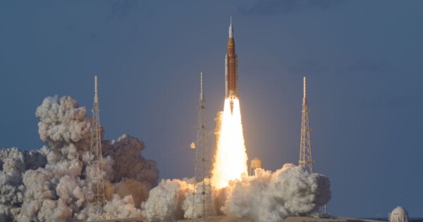 Foguete sendo lançado a partir de uma base de lançamento ao ar livre. A imagem mostra o momento inicial da decolagem, com uma grande chama intensa saindo da base do foguete e uma nuvem densa de fumaça branca e cinza se espalhando ao redor. Estruturas metálicas altas cercam a plataforma de lançamento. O céu ao fundo é azul claro, sem nuvens, e a iluminação natural destaca o contraste entre o fogo brilhante e a fumaça. (SpaceX